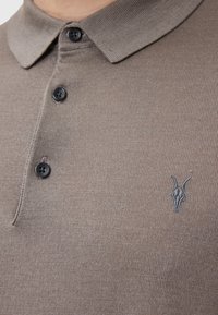 Brun stickad polotröja med krage, försedda med tre svarta knappar och en grå getskallebroderi på vänster bröst.