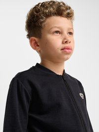 Dreng med krøllet hår iført en sort lynlåstrøje med et lille hvidt logo, der kigger let op og til siden mod en ensfarvet baggrund.