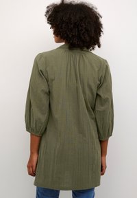 Tunika verde oliva, strutturata, con maniche a tre quarti, leggermente arricciata sul retro e dal taglio rilassato, abbinata a jeans blu.