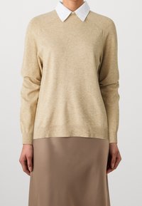 Beżowy sweter dzianinowy z białą koszulą z kołnierzykiem pod spodem; ma długie rękawy, prążkowany dół i subtelną teksturę. W zestawieniu z brązową spódnicą.