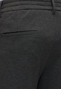 Détail de la poche arrière et de la ceinture sur un pantalon gris foncé sur mesure, avec une boucle de ceinture visible sur le côté droit.