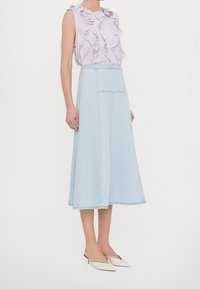 Kvinna iklädd ärmlös lavendelfärgad blus med volanger, ljusblå högmidjad midi denimkjol och spetsiga vita låga klackskor, stående på sidan.