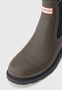 Botte de cheville en caoutchouc vert foncé avec des côtés élastiques noirs, présentant un talon texturé, un capuchon de pied lisse et un logo "HUNTER" rouge et blanc.