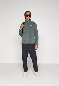 Fleecejakke i dempet grønn, glidelås foran, høy krage, kombinert med svarte bukser og beige joggesko, modell som holder en ryggsekk, iført solbriller.