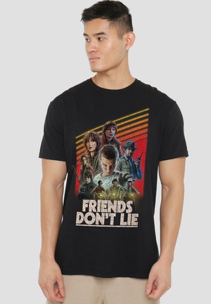 Junger Mann trägt ein schwarzes T-Shirt mit Charakteren aus Stranger Things und dem Text "Freunde lügen nicht" in fetten Buchstaben.