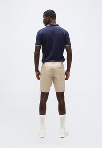 Άνδρας φορώντας μπλε ναυτικό κοντομάνικο πόλο, μπεζ σορτς, λευκές κάλτσες μέχρι τον αστράγαλο και λευκά αθλητικά παπούτσια, στέκεται με την πλάτη προς το μέρος μας σε απλό φόντο.