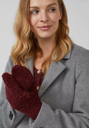 Frau mit welligem blonden Haar, die einen grauen Mantel und einen kastanienbraunen Strickcardigan trägt, hält passende kastanienbraune strukturierte Handschuhe nahe ihr Gesicht.