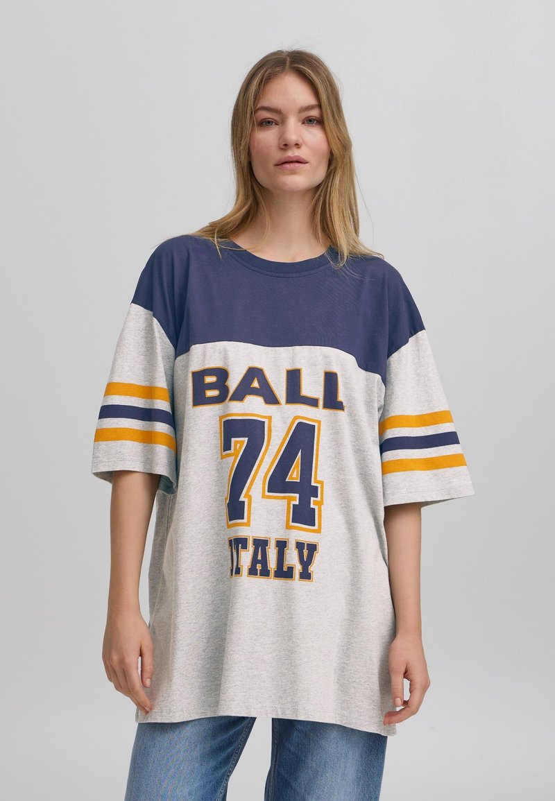 Tricou oversized gri cu dungi albastre marine și galbene, având înscrisul "BALL 74 ITALY" tipărit cu litere mari pe față.