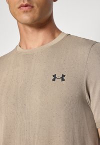 Beige crew-neck t-skjorte laget av teksturert stoff med svake vertikale striper. Har en svart logo på venstre bryst.