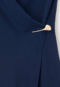 Tissu bleu marine plié et maintenu par une épingle métallique lisse dorée formant un motif de feuille minimaliste.