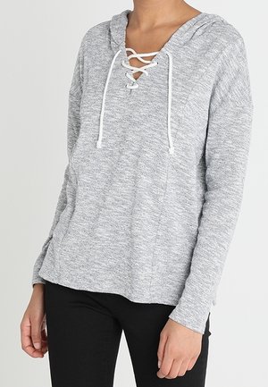 Hellgraues langärmliges Kapuzenshirt mit weißem Schnürdetail am Ausschnitt, getragen von einer stehenden Person, kombiniert mit schwarzen Hosen.