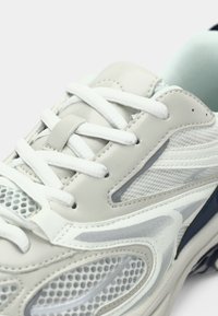 Ανοιχτό γκρι αθλητικό παπούτσι με πάνελ από δίχτυ, λευκά κορδόνια και ναυτικό μπλε λεπτομέρεια· διαθέτει επένδυση στον γιακά και ξεχωριστό σχεδιασμό επικάλυψης.