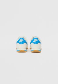 Baltas un zils Nike apavu pāris ar teksturētu gumijas zoli, noapaļotu formu un logotipa akcentiem uz papēža un sāniem.