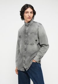 Grijze denim shirt met lange mouwen, knopen aan de voorkant, borstzak en een licht vervaagde textuur. Model draagt donkerblauwe spijkerbroek.