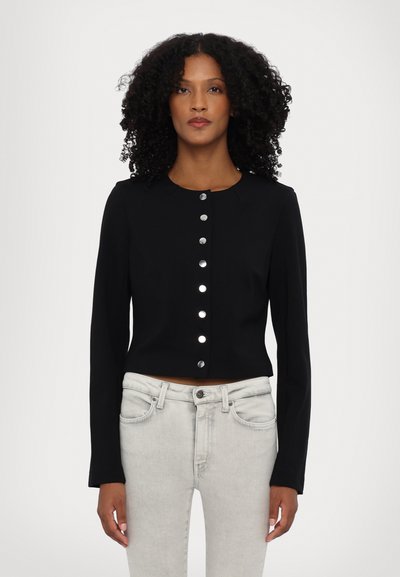 Veste noire courte à manches longues avec des boutons-pression argentés sur le devant, portée sur un jean gris clair. Texture de tissu lisse.