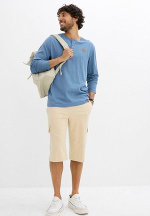 Maglietta henley a maniche lunghe azzurro chiaro, pantaloni cargo beige e sneakers bianche. La maglietta presenta dettagli con bottoni e una piccola etichetta in pelle.