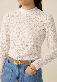 Weißes Spitzenoberteil mit langen Ärmeln und hohem Kragen, florales Muster, filigrane Details, kombiniert mit einem blauen Jeansrock und einem hellbraunen Gürtel.