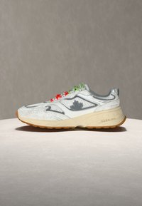 Baskets blanches et grises avec une semelle beige, des lacets rouges et verts, et un logo de feuille d'érable grise sur le côté, présentées sur une surface ronde et claire.