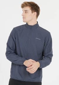 Tummansininen kevyt takki, jossa on pystykaulus, vetoketjukiinnitys, elastiset hihansuut ja huomaamaton logotyyli rinnassa.