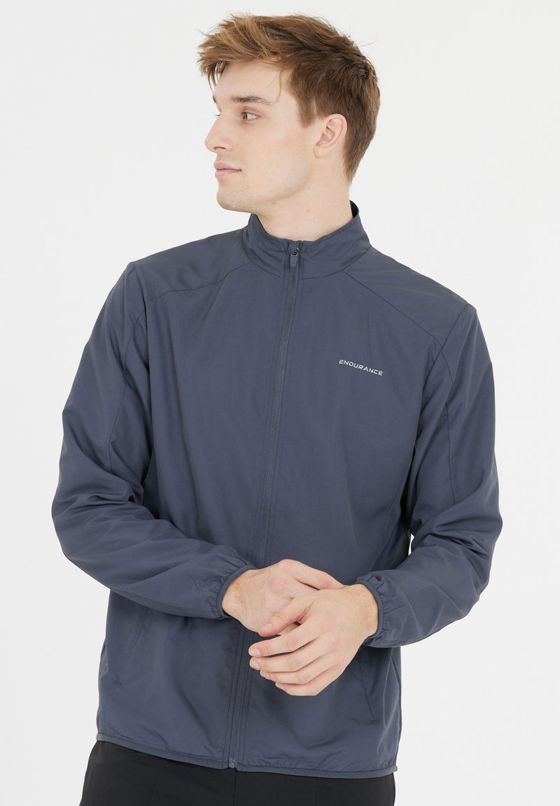 Tummansininen kevyt takki, jossa on pystykaulus, vetoketjukiinnitys, elastiset hihansuut ja huomaamaton logotyyli rinnassa.