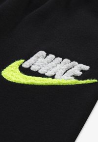 A fekete anyagon egy feltűnő hímzett Nike logó található fehér színben, élénk sárgászöld árnyalatokkal, megjelenítve a texturált varrás részleteit.