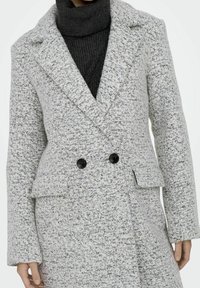 Žena v šedém skvrnitém dvouřadém vlněném kabátu s černými knoflíky, obléká tmavě šedý rolák, ruce uvolněně položené po stranách.