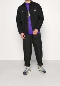 Uomo che indossa una giacca nera con bottoni, un maglione grafico viola, pantaloni cargo neri e sneaker bianco-grigi che sta in piedi su un pavimento grigio.