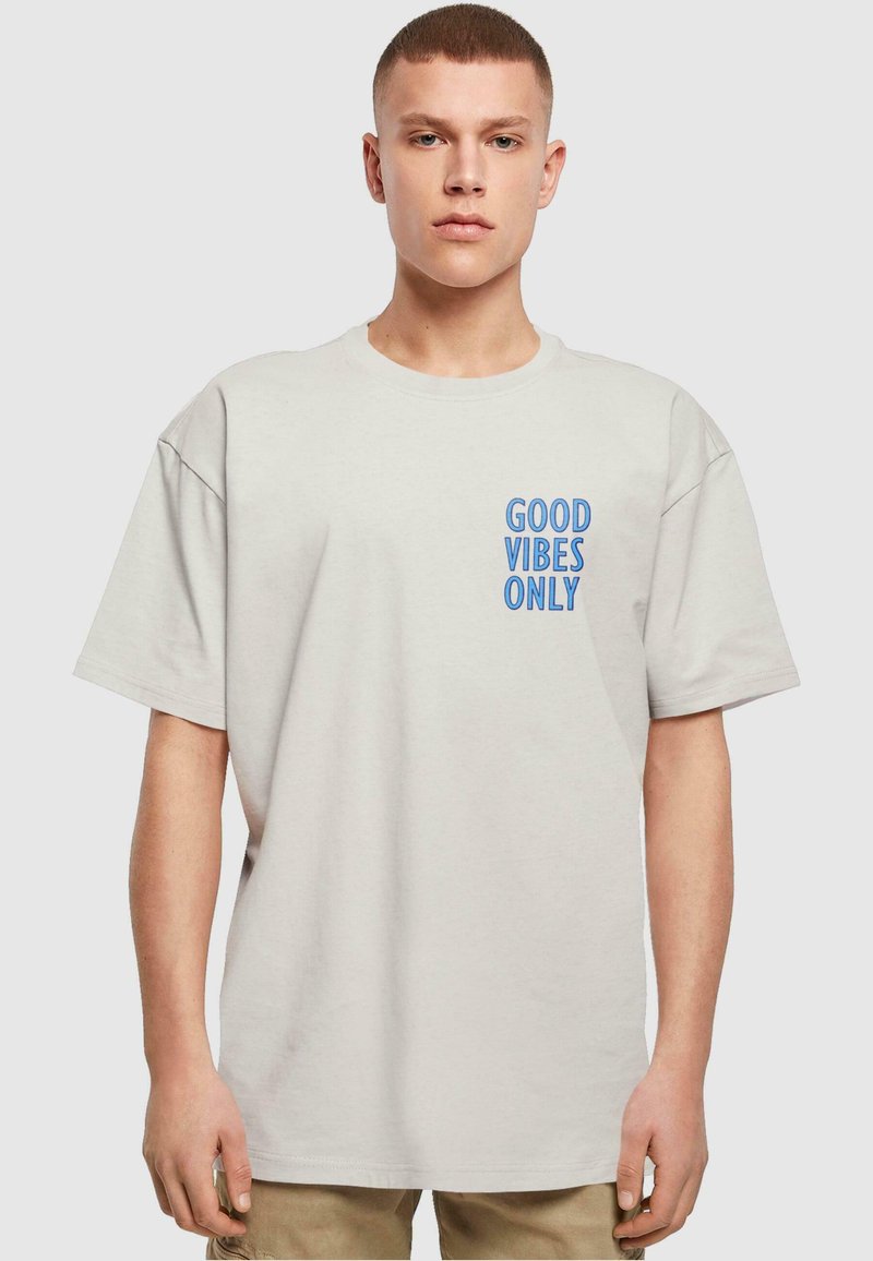 Lichtgrijs katoenen t-shirt met een blauwe graphic die "GOOD VIBES ONLY" op de linkerkant van de borst leest. Losse pasvorm met korte mouwen.