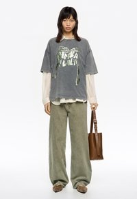 Camiseta gris de gran tamaño con texto gráfico verde, combinada con pantalones sueltos de color verde oliva y un bolso de piel marrón.