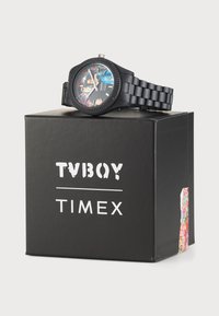 Black plastic wristwatch with a multi-colored face, featuring a textured bezel and a patterned strap, placed on a black box with white text.