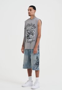 Camiseta gráfica sin mangas gris con estampado negro, combinada con shorts de mezclilla azul claro que presentan acentos de calavera y diseño en negro. Zapatillas blancas completan el look.