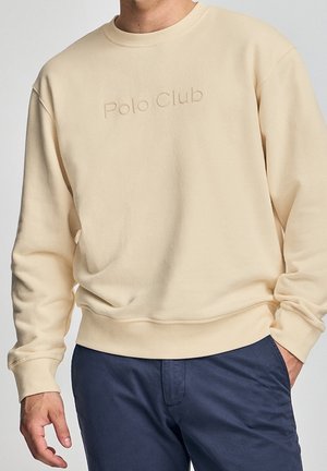 Man draagt beige sweatshirt met verdiept 'Polo Club' tekst en donkerblauwe broek, handen deels zichtbaar, effen achtergrond.