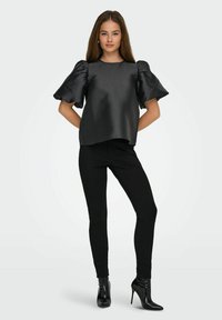 Joven mujer vestida con una blusa de satén negra de mangas abullonadas, pantalones negros ajustados con detalles laterales y botines de tacón alto negros, de pie con confianza.