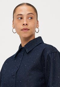 Donkerblauwe denim shirt met een klassieke kraag, voorzien van metalen studs verspreid over de stof, wat textuur en ontwerp accentueert.