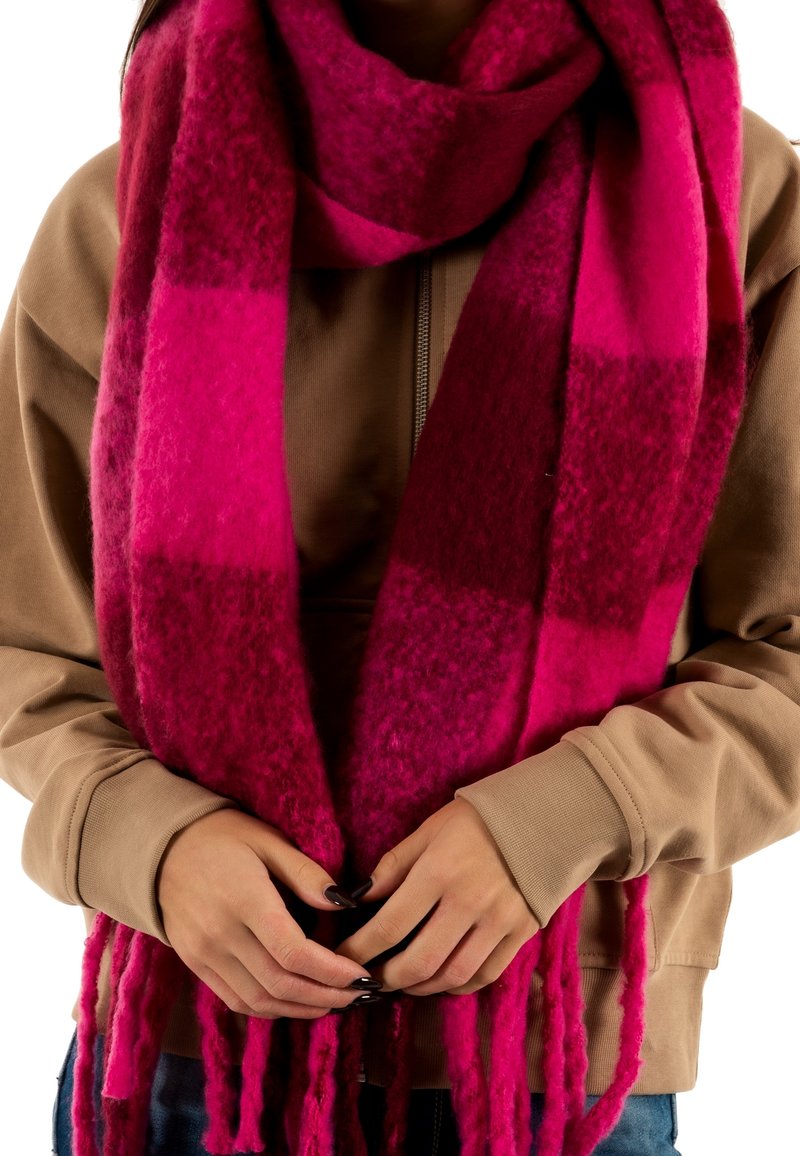 Écharpe à carreaux fuchsia en tissu épais et doux, avec des extrémités frangées ; superposée à un sweat à capuche marron, les mains tenant l'écharpe.