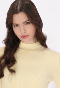 Maglione a maniche lunghe con collo a dolcevita in morbido tessuto giallo. Caratterizzato da una forma aderente e una leggera trama. I capelli sono mossi e castano scuro.