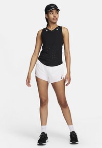 Juodas be rankovių sportinis marškinėlis su perforuotu dizainu, derinamas su baltais šortais. Aksesuarai: juoda kepurė ir juodi sportbačiai.