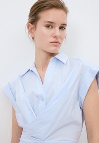 Vaaleansininen raidallinen paita, jossa on kääntömalli, lyhyet hihat ja terävä kaulus. Pehmeä kangas, jossa on nappikiinnitys kaula-aukossa.