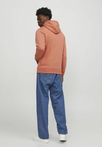 Sudadera con capucha en suave color coral, combinada con jeans azules sueltos que cuentan con una tela texturizada y un detalle de etiqueta en el lateral, usada con zapatillas blancas.