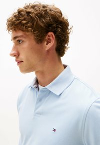 Jasnoniebieska koszulka polo z teksturowanej tkaniny, z kołnierzykiem i małym haftowanym logo w czerwonym, białym i niebieskim kolorze na piersi.