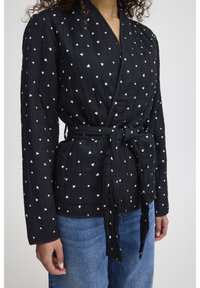 Veste noire enveloppante avec des motifs de cœurs blancs, manches longues, tissu texturé et une ceinture à nouer à la taille. Portée par-dessus un pantalon en denim bleu.