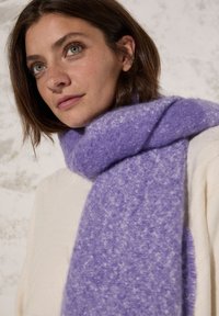 Écharpe tricotée violette en matériau doux, avec une finition texturée et des variations de couleur subtiles, drapée autour du cou.