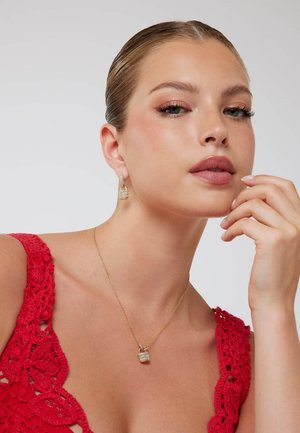 Collana d'oro con ciondolo a lucchetto piccolo e orecchini con design a lucchetto abbinato. Modella indossa una maglia rossa di pizzo. Texture della pelle liscia.