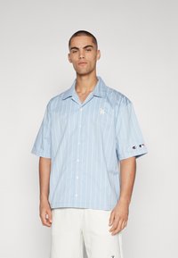 Camisa de manga curta azul clara com riscas verticais brancas. Apresenta um fecho frontal com botões, colarinho pontiagudo e logótipo bordado no lado esquerdo do peito.
