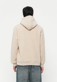 Mann in einer hellbeigen Fleece-Haube und lockeren blauen Jeans, der mit dem Rücken zu einem schlichten weißen Hintergrund steht.