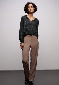 Tummanvihreä pusero, jossa on pieniä valkoisia täpliä, v-aukkoinen kaula-aukko ja elastiset hihansuusuut, yhdistettynä vaaleanruskeisiin, räätälöityihin housuihin ja mustiin kenkiin.