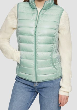 Nainen pukeutunut vaaleanvihreään tikattuun hihattomaan liiviin kermanvärisen neulepuseron ja sinisten farkkujen päällä, seisomassa käsi taskussa.