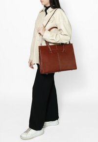 Borsa a tracolla in pelle marrone con doppio manico superiore, superficie liscia e dettaglio con cucitura centrale. Indossata con un giubbotto color crema e pantaloni neri.