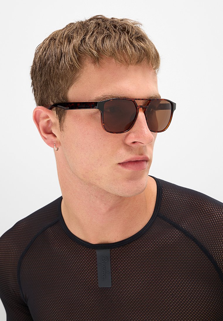 Młody mężczyzna z krótkimi, jasnobrązowymi włosami, noszący okulary przeciwsłoneczne w tygrysie cętki, mały kolczyk włożony w uchu i czarny top z siateczki z metką Rapha.