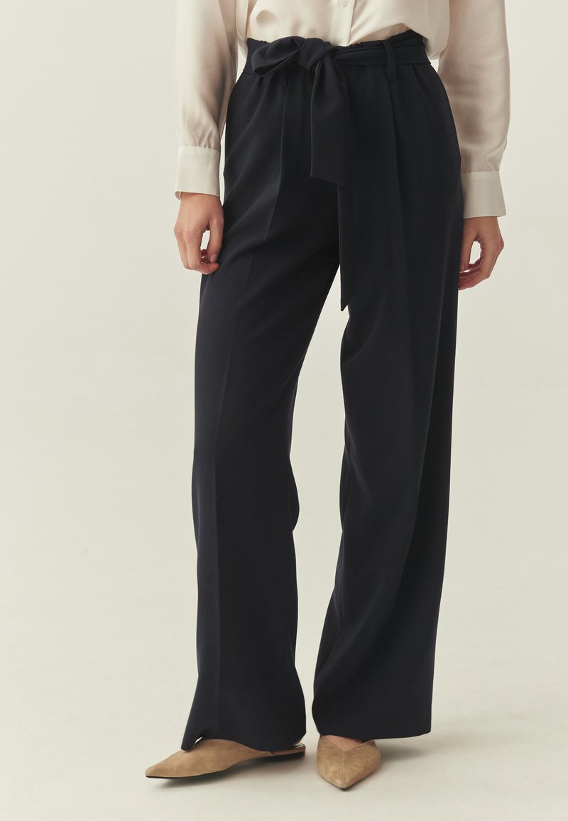 Pantalon large bleu marine avec ceinture en tissu, présentant une texture lisse et des détails plissés. Associé à des chaussures de couleur claire.